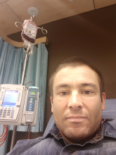A man from Australia sits close to the camera in his hospital room. An I-V system is in the photo behind him, with a bag of blood product attached.