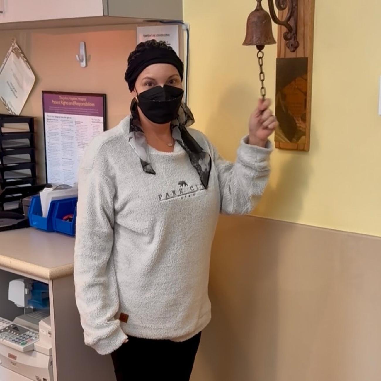 Photo of Kelsey at the end of treatment. She is standing next to the bell for patients to ring at the end of treatment. She is wearing a sweatshirt and pants, along with a mask and a scarf.